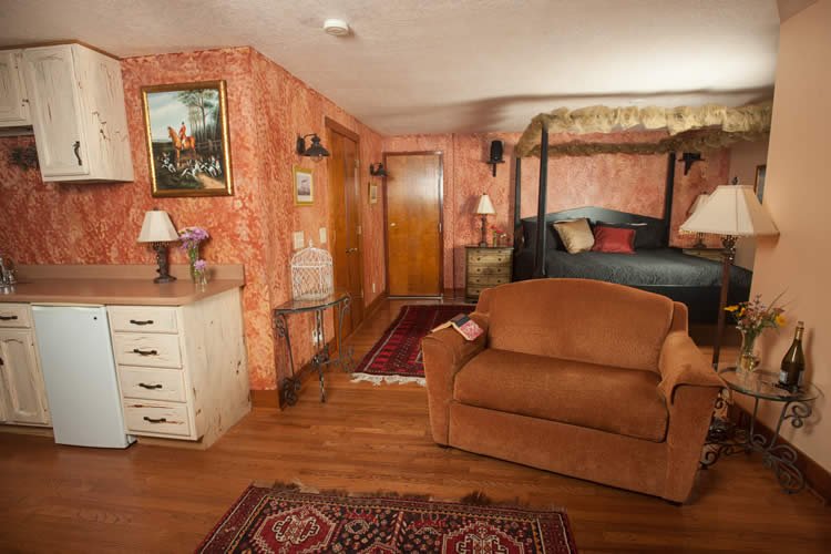 Room with canopy bed, dark bedspread, Kitchenette area with refrig and sink, room done in peach color and hardwood floor